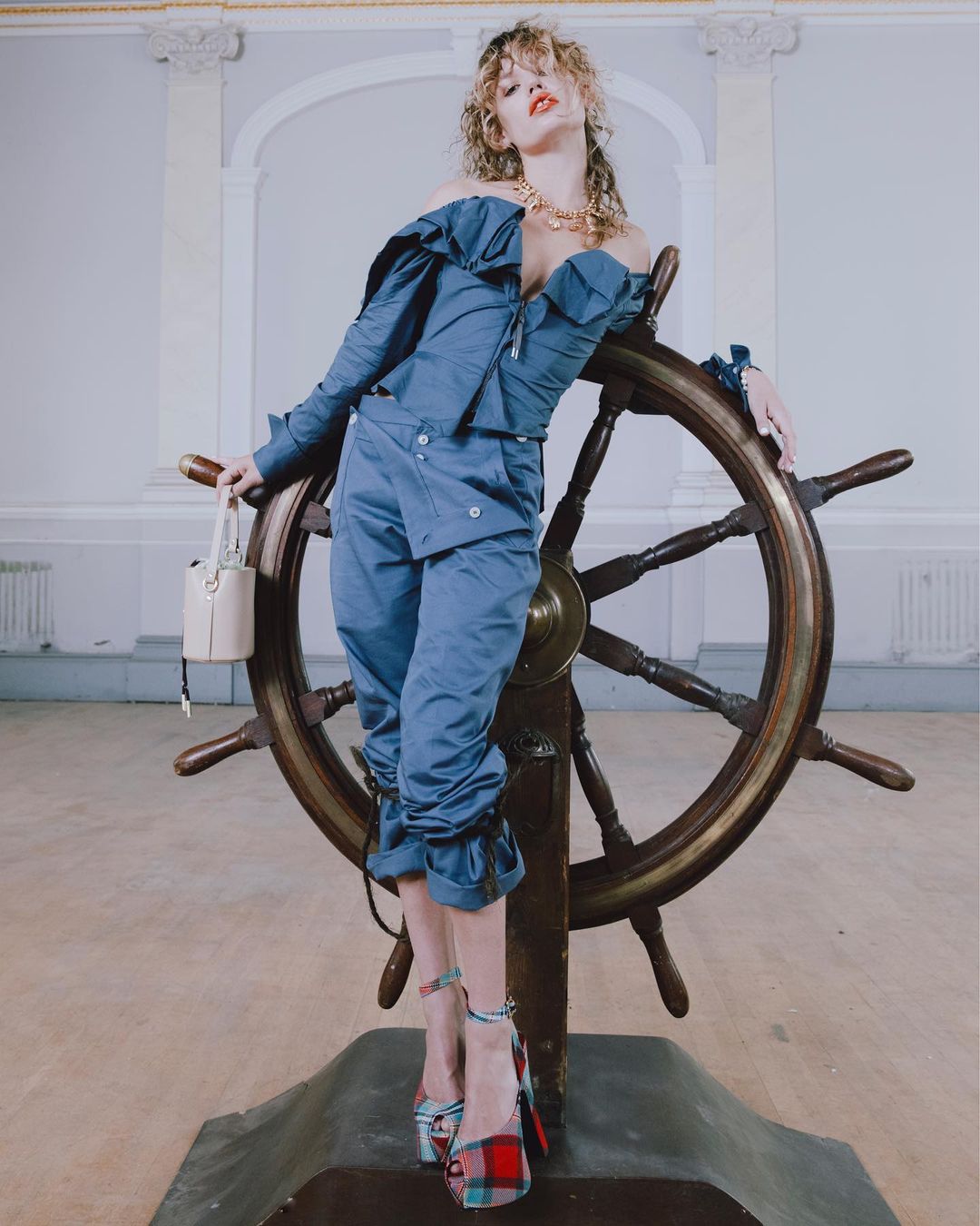 A blonde woman in a blue off-the-shoulder outfit poses with a ship's wheel in a light-filled interior, wearing platform shoes and holding a small bucket bag.