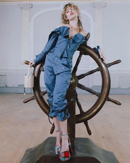 A blonde woman in a blue off-the-shoulder outfit poses with a ship's wheel in a light-filled interior, wearing platform shoes and holding a small bucket bag.