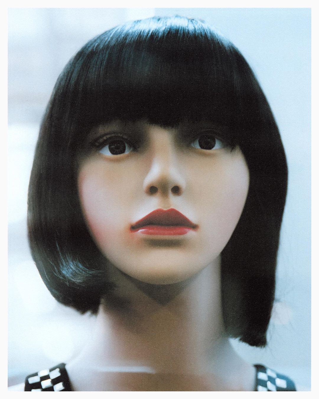 Portrait of a mannequin with black bob haircut, red lipstick, and checkered top against a pale blue studio background with even lighting.