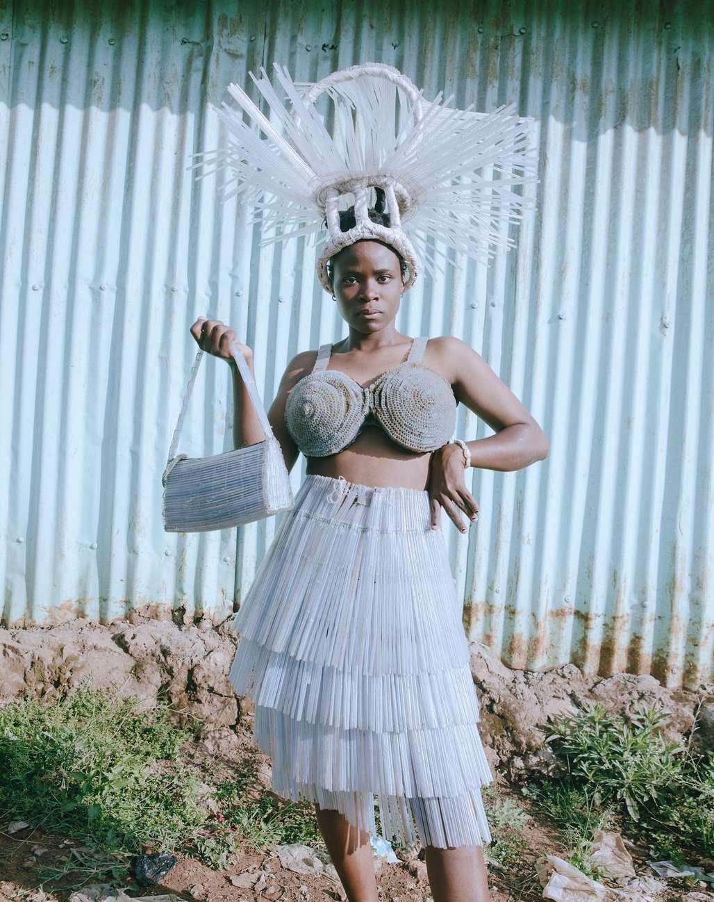 Woman in upcycled fashion poses against corrugated metal wall, wearing sculptural headdress, bustier top, and tiered skirt.