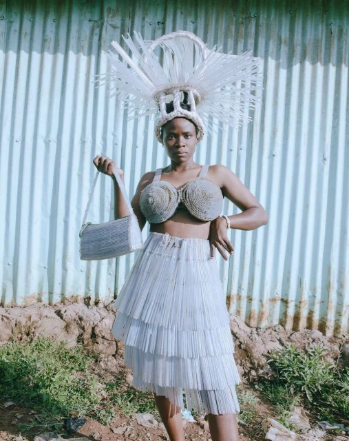 Woman in upcycled fashion poses against corrugated metal wall, wearing sculptural headdress, bustier top, and tiered skirt.