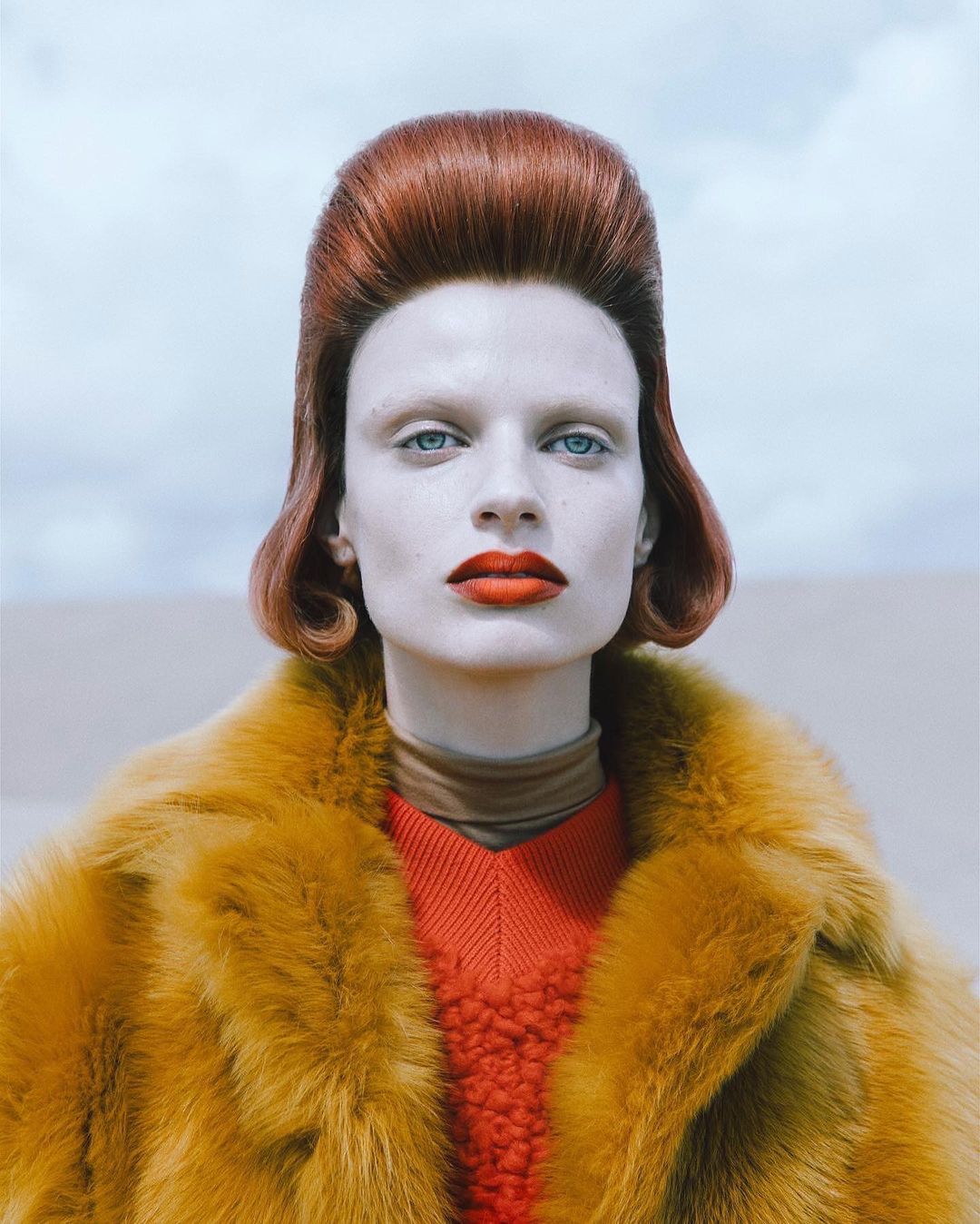 Fashion shot of a woman with red hair and red lipstick wearing a golden fur coat in front of a light blue sky and a light beige wall.