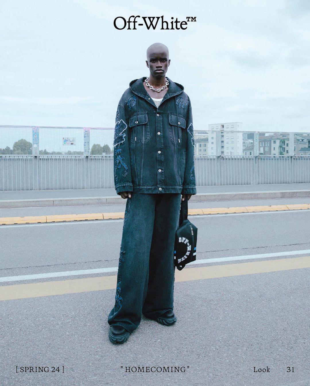Black male model wearing dark distressed denim jacket and matching wide leg pants standing on asphalt road with bridge background