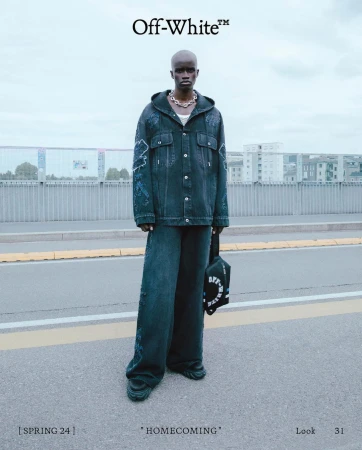 Black male model wearing dark distressed denim jacket and matching wide leg pants standing on asphalt road with bridge background