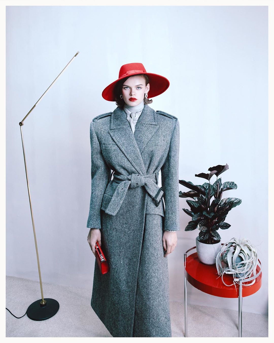 Fashion portrait of an adult woman in a grey coat and red hat, holding a red clutch, posed in a photo studio with modern decor.