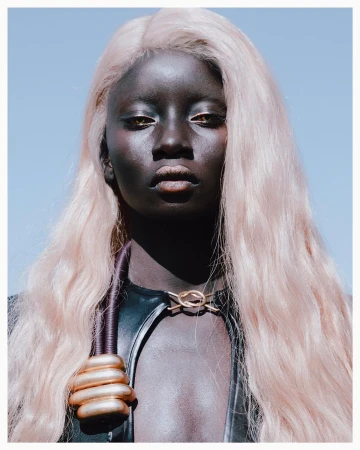 Portrait of a Black woman with long pink hair and gold metallic neckwear against a clear blue sky by Kristin-lee Moolman