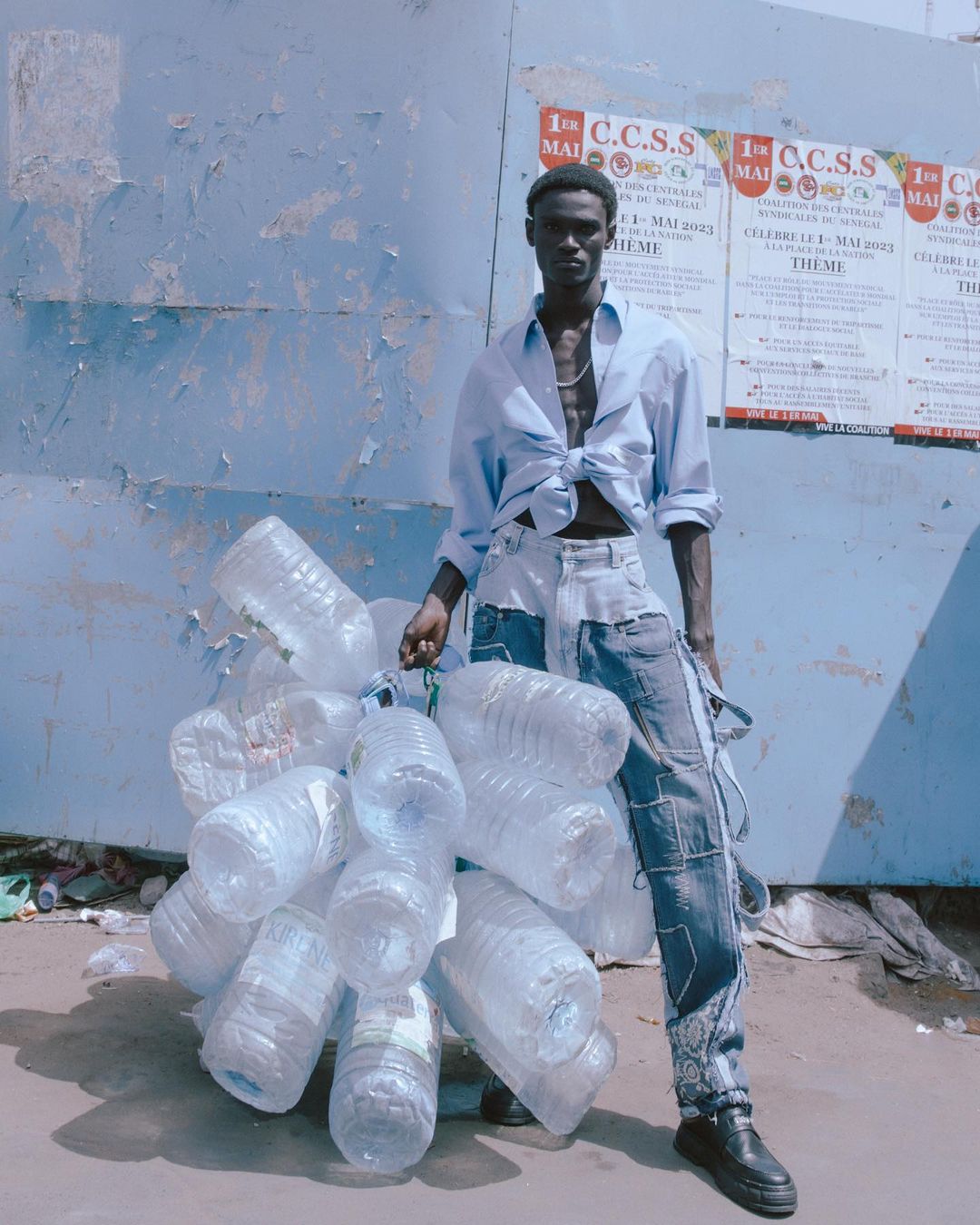 A slender man strikes a pose outdoors, wearing a light blue tied shirt and patchwork jeans, holding a collection of empty plastic bottles.