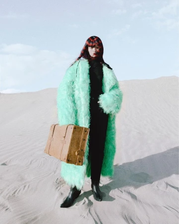 A young female model in a light green faux fur coat stands in a desert carrying a vintage suitcase under a blue sky with light clouds.
