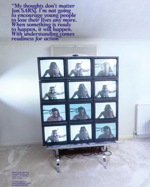 Installation art of a stack of old CRT TVs showing a shirtless man with dreadlocks inside a minimal, naturally-lit interior.