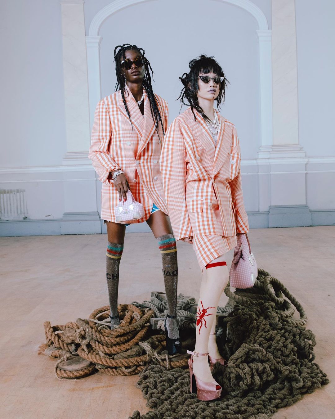 Two fashion models in matching plaid blazers pose indoors on a pile of rope with light pink purses and platform heels in a fashion editorial.