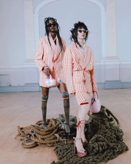 Two fashion models in matching plaid blazers pose indoors on a pile of rope with light pink purses and platform heels in a fashion editorial.