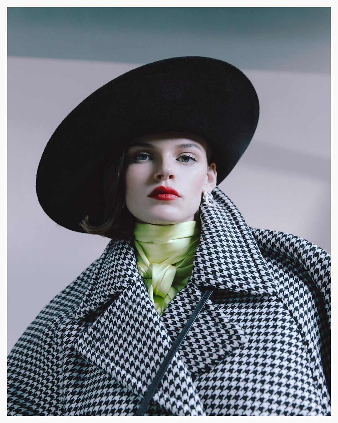 Elegant woman in wide-brimmed black hat and houndstooth coat poses indoors with a light yellow top and red lipstick in a fashion shot.
