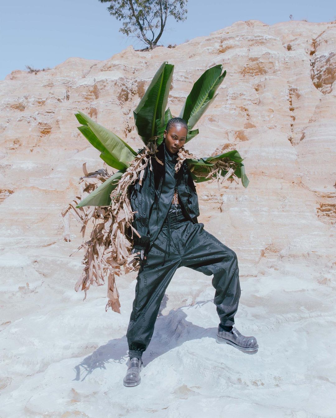 A striking fashion shot of a woman in a black jumpsuit, holding banana leaves, posing in front of a light brown rock formation.