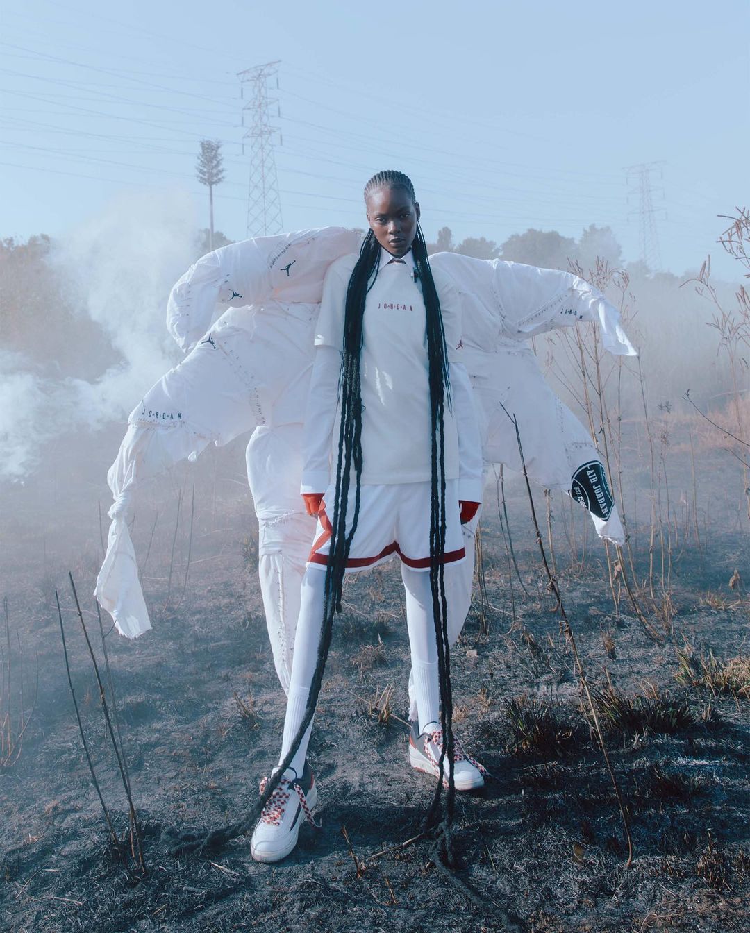 Editorial fashion shot of a woman wearing a white Jordan outfit and long braids, posing outdoors in a field with smoke and powerlines.