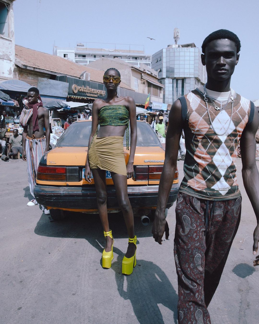 Three models pose on a city street in Senegal with an old yellow car, wearing trendy outfits and statement platform shoes.