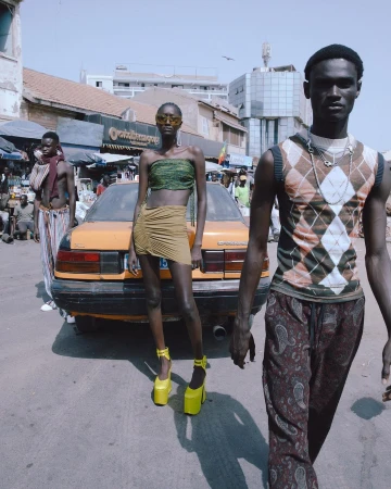 Three models pose on a city street in Senegal with an old yellow car, wearing trendy outfits and statement platform shoes.