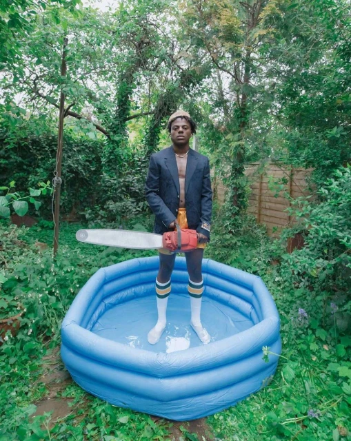 Adult male stands in a blue kiddie pool in backyard, wearing a blazer, shorts, socks, and holding a chainsaw. Green foliage backdrop.