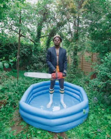Adult male stands in a blue kiddie pool in backyard, wearing a blazer, shorts, socks, and holding a chainsaw. Green foliage backdrop.