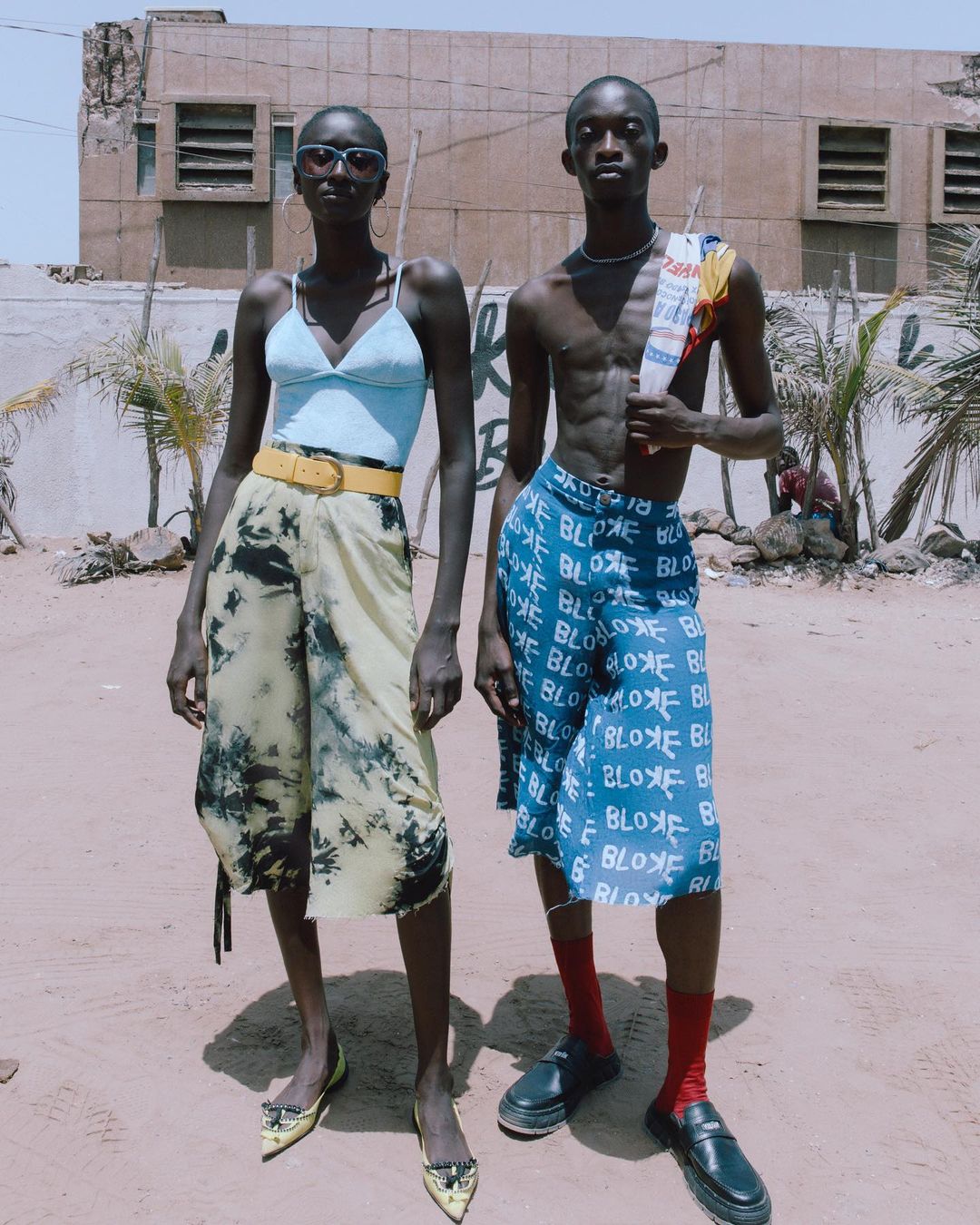 Two fashion models pose outdoors wearing light blue tops and knee-length bottoms with yellow and black shoes and red socks in the sun.