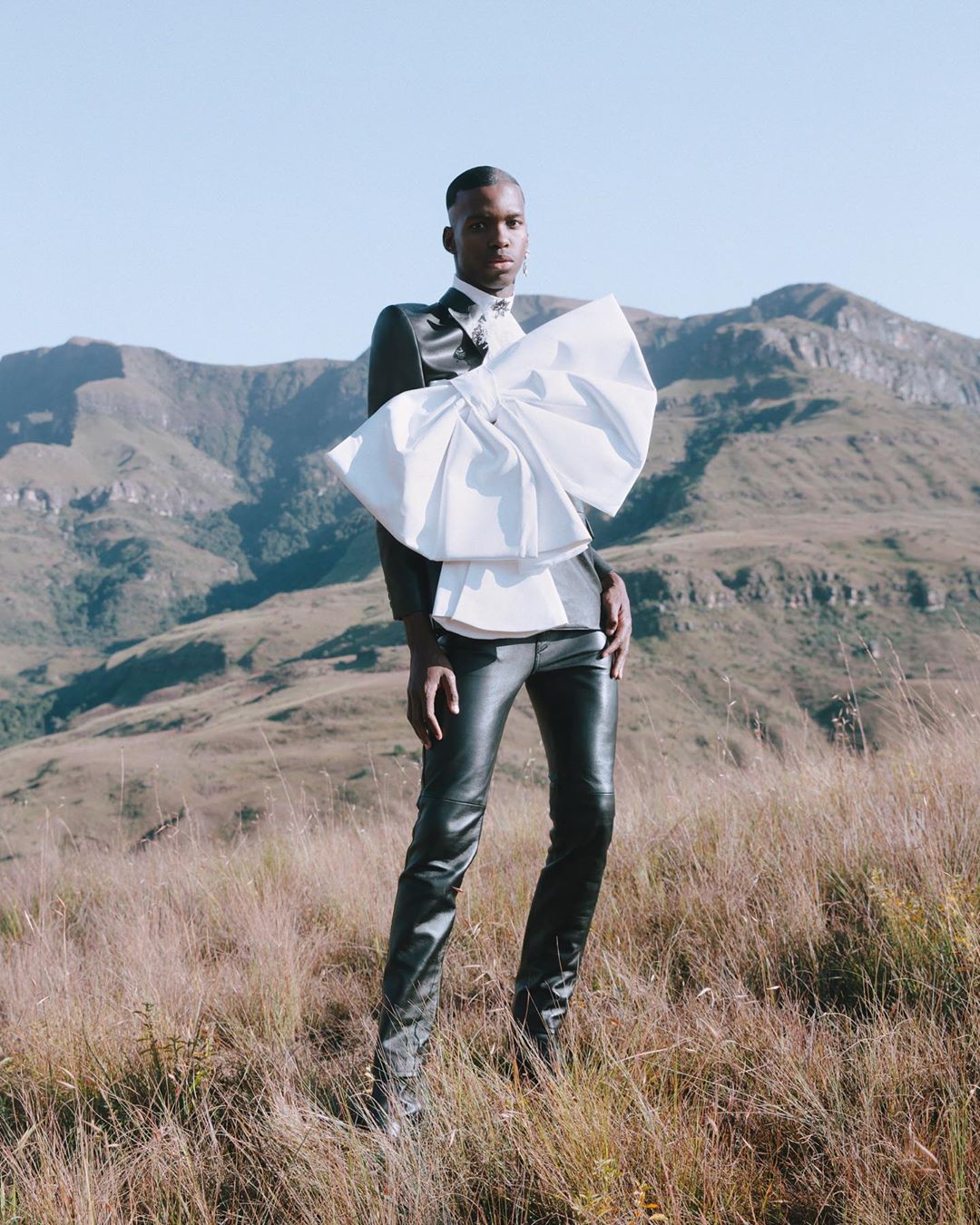 Adult male fashion model posing in a black jacket, shiny black pants, and a large white bow outside on a grassy hillside.