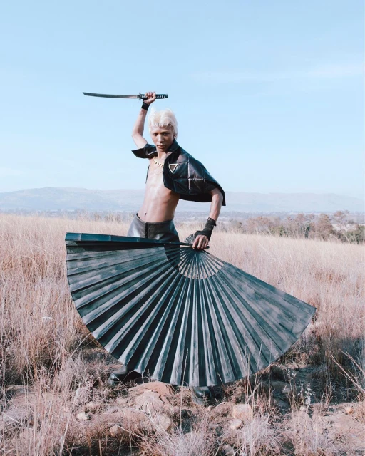 A person poses outdoors holding a katana sword and a folding fan, wearing a black vest, with a grassy landscape and blue sky backdrop.