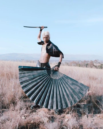 A person poses outdoors holding a katana sword and a folding fan, wearing a black vest, with a grassy landscape and blue sky backdrop.