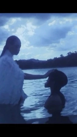 Two people stand in water against a sky with clouds, one adult places a hand on a child's head in a tranquil scene.