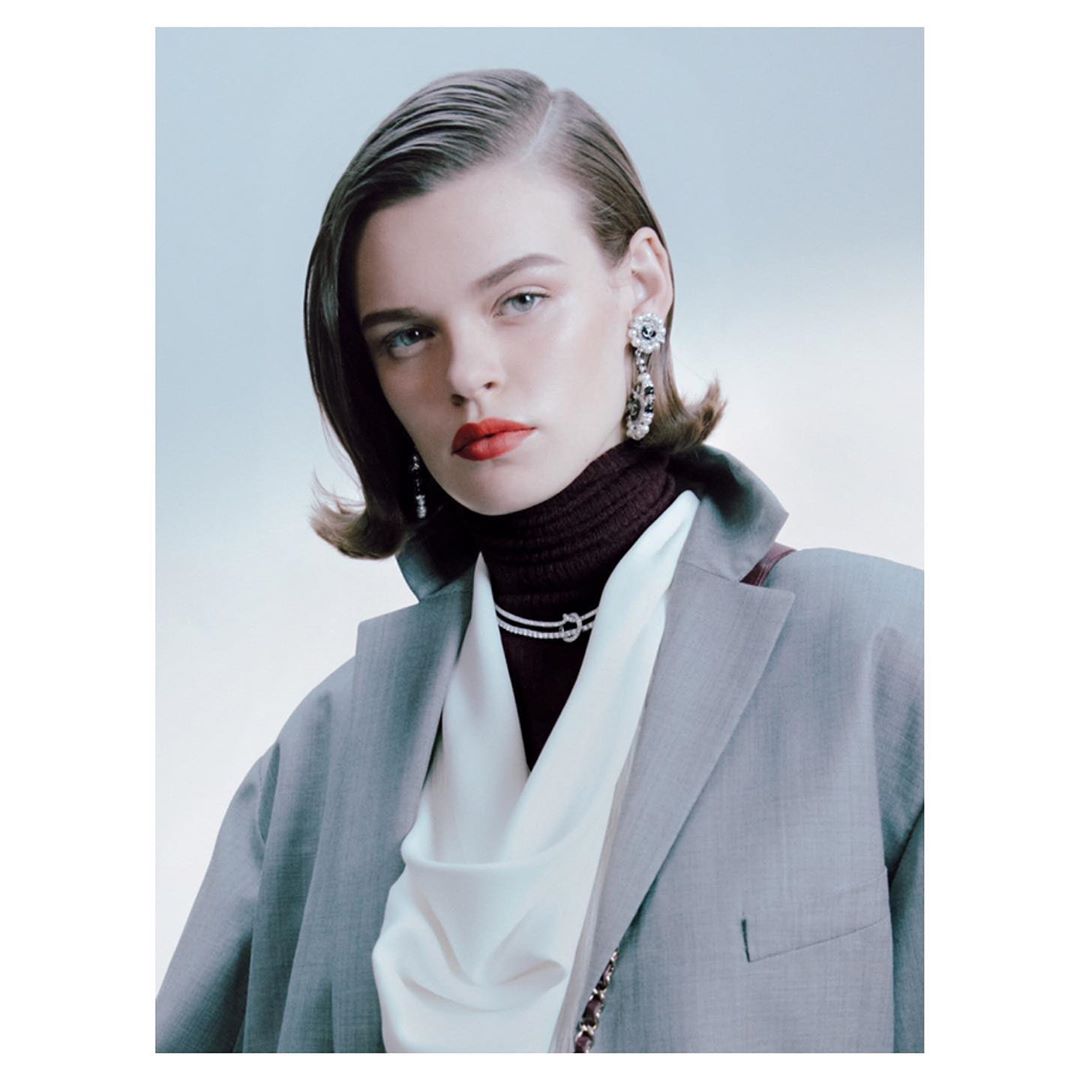 Portrait of a stylish woman with dark blonde hair, wearing a gray blazer, turtleneck, jewelry, and red lipstick against a light background.