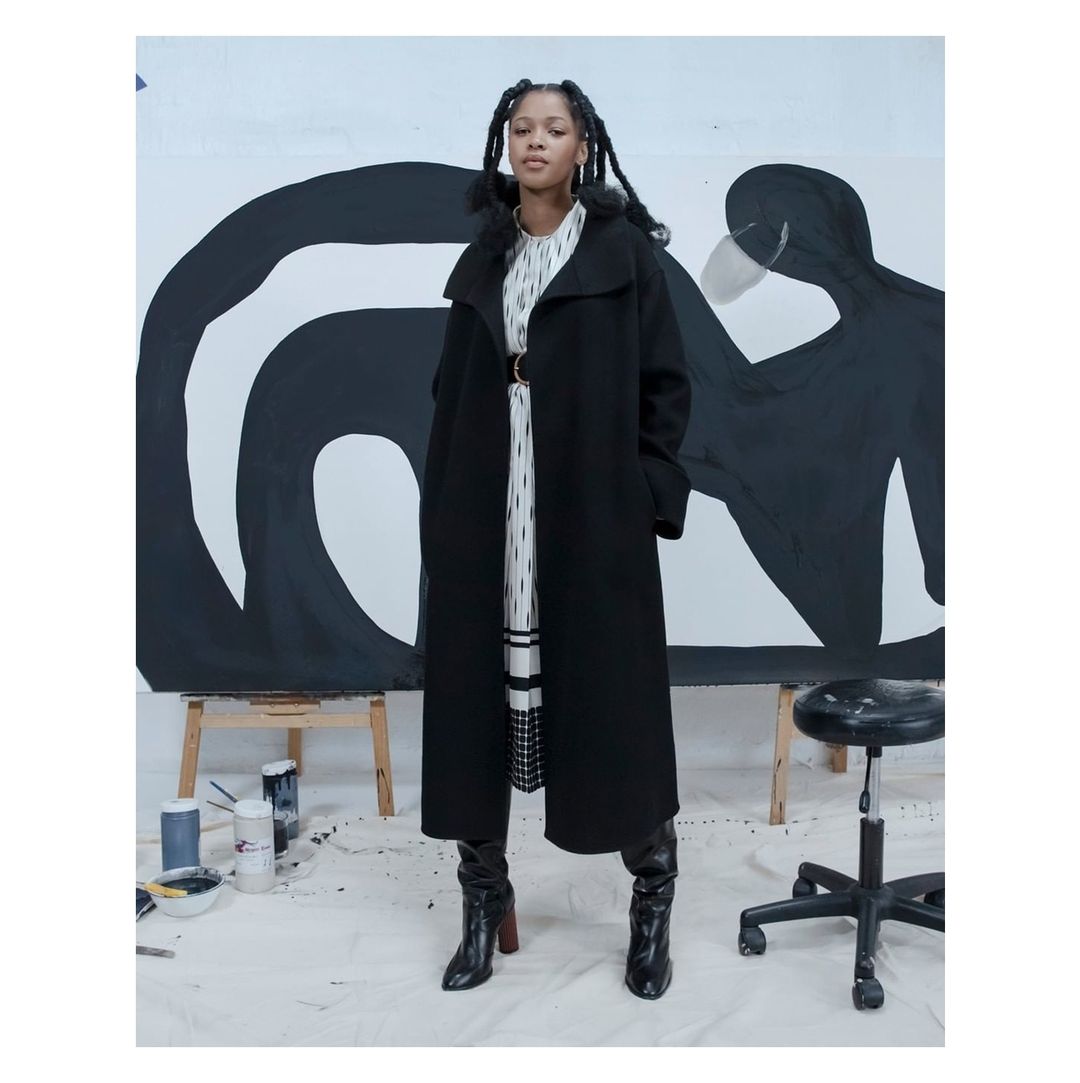 A Black woman in a long black coat poses in a photo studio against a background of black-and-white abstract painting.