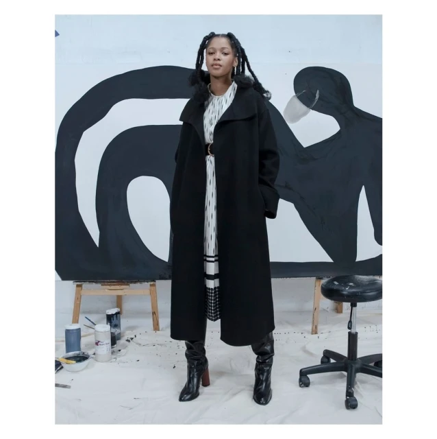 A Black woman in a long black coat poses in a photo studio against a background of black-and-white abstract painting.