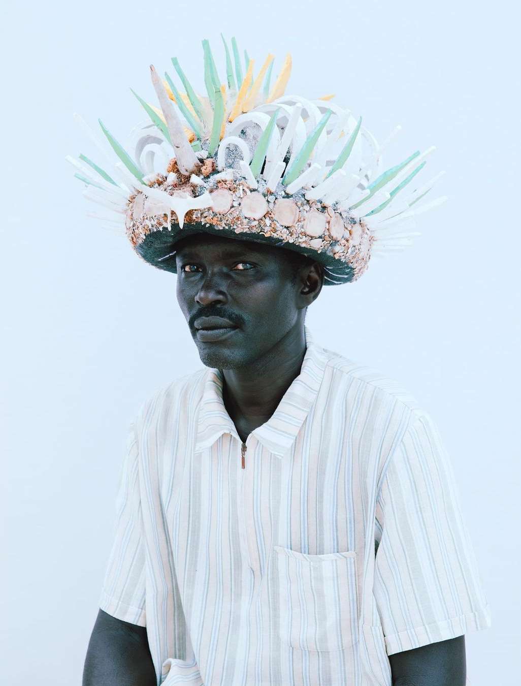 Adult male posing in a photo studio with a decorative headdress made of shells and colorful plastic, wearing a striped shirt.