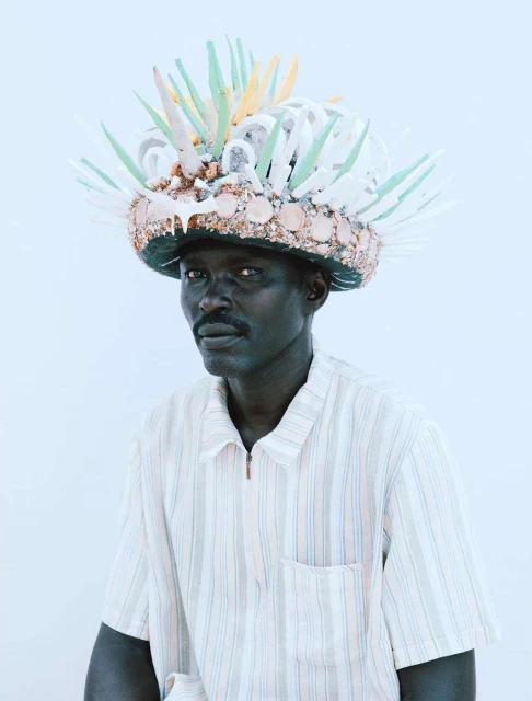 Adult male posing in a photo studio with a decorative headdress made of shells and colorful plastic, wearing a striped shirt.