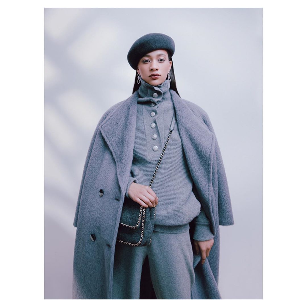 Fashionable young woman in grey wool coat, beret, sweater, and pants, carrying a chain-strap bag against a neutral backdrop.