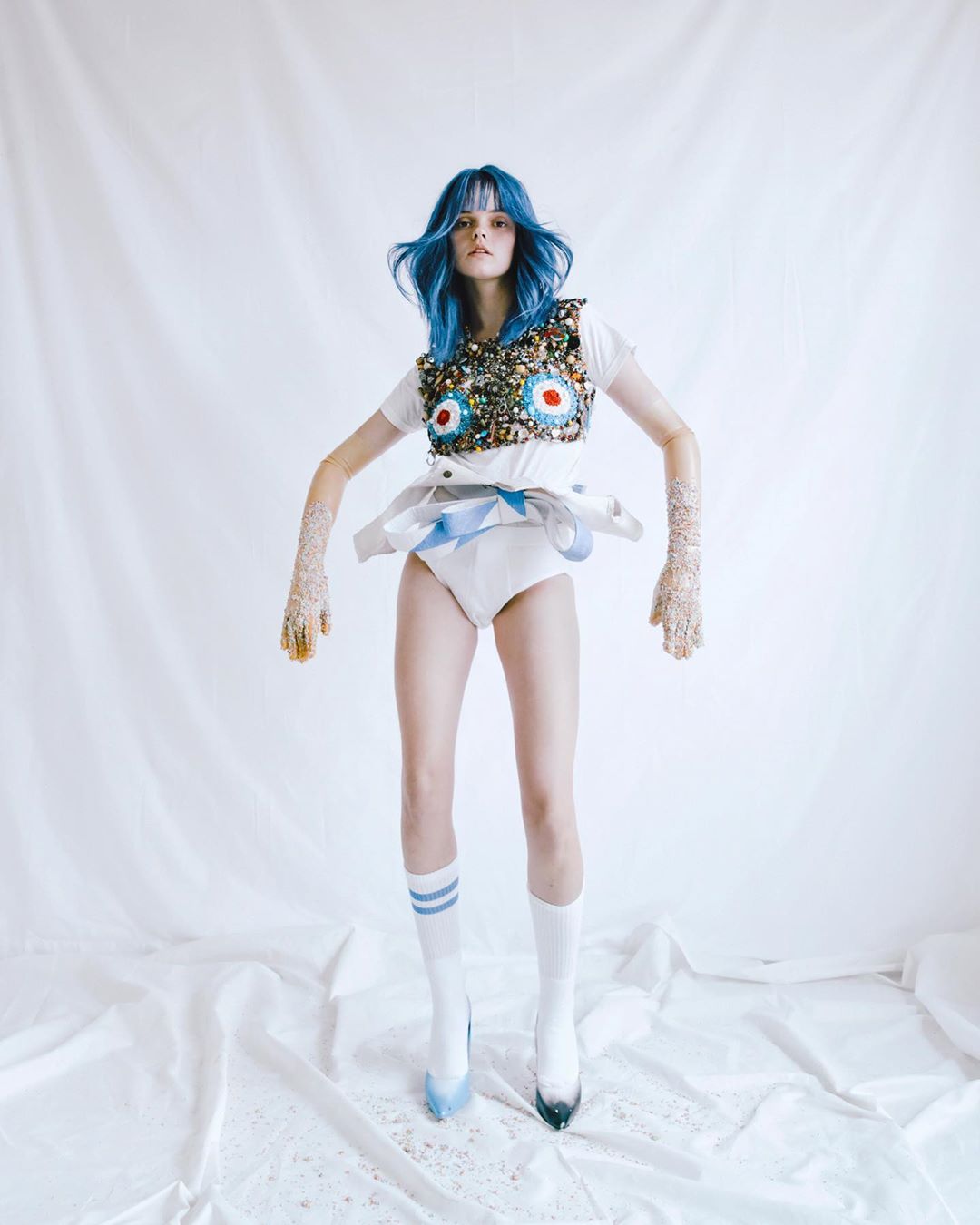 Woman with blue hair and an embellished top jumps in a white room. The photograph is a fashion portrait, highlighting textures and materials.
