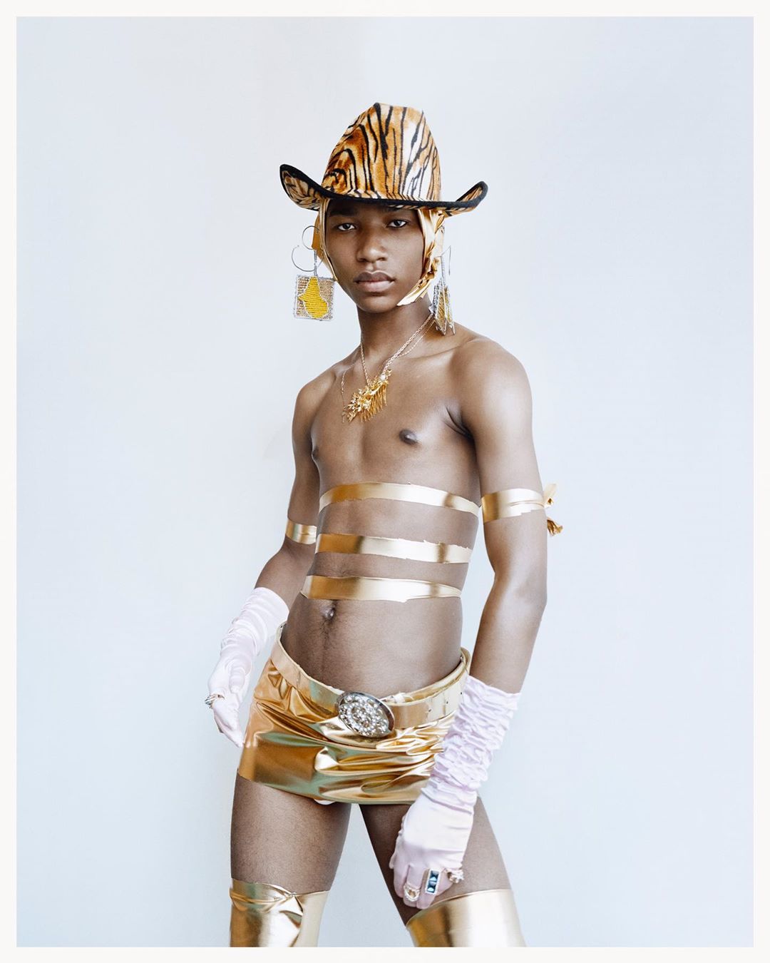 Person wearing a tiger-print hat, gold-colored top, skirt, and boots poses against a white wall, accentuated by gold armbands and jewelry.