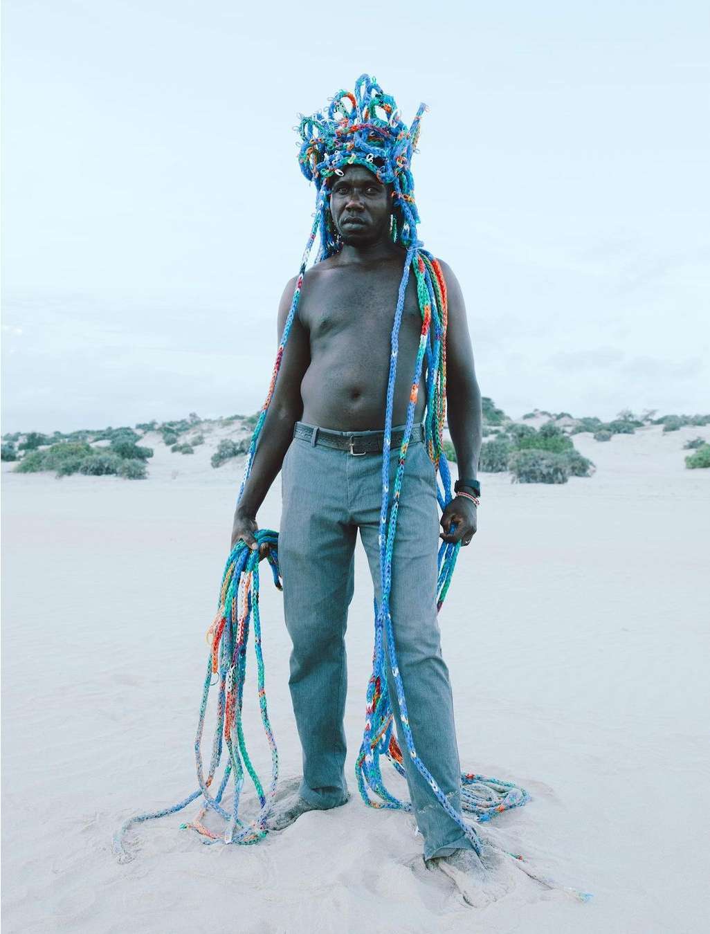 Person stands on sandy beach wearing an elaborate colorful headdress and draped with matching strands of colorful rope.