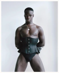 Black man wearing a black corset and silver pendant necklace against a plain white studio background