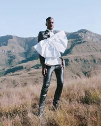 Black man standing in dry grass wearing a black leather jacket and large white bow against a mountain backdrop