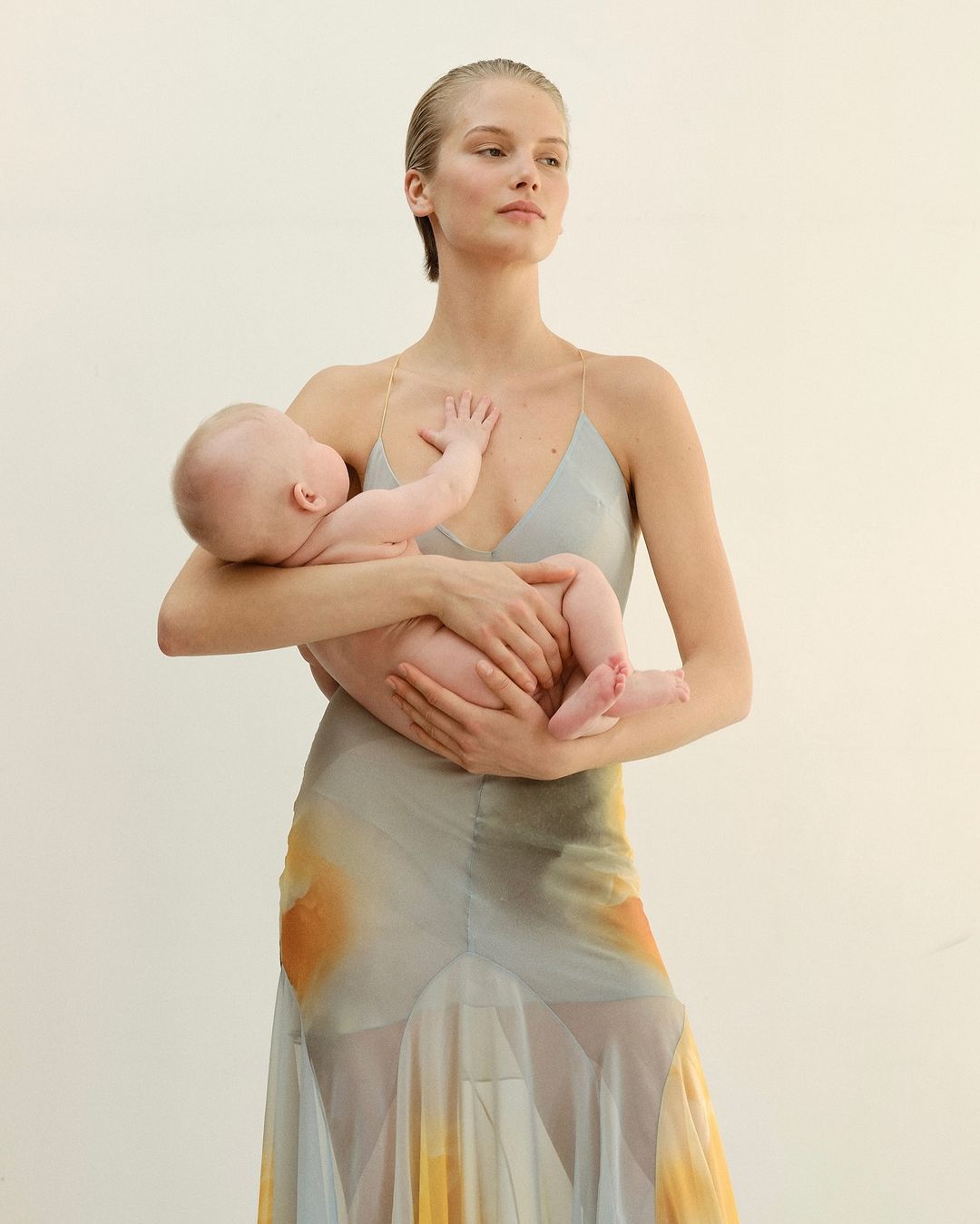 Woman with slicked-back hair holding an infant while wearing a patterned slip dress against white seamless backdrop