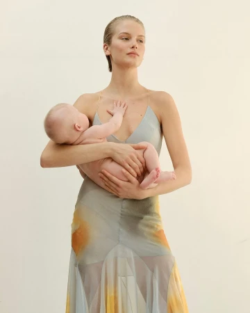 Woman with slicked-back hair holding an infant while wearing a patterned slip dress against white seamless backdrop