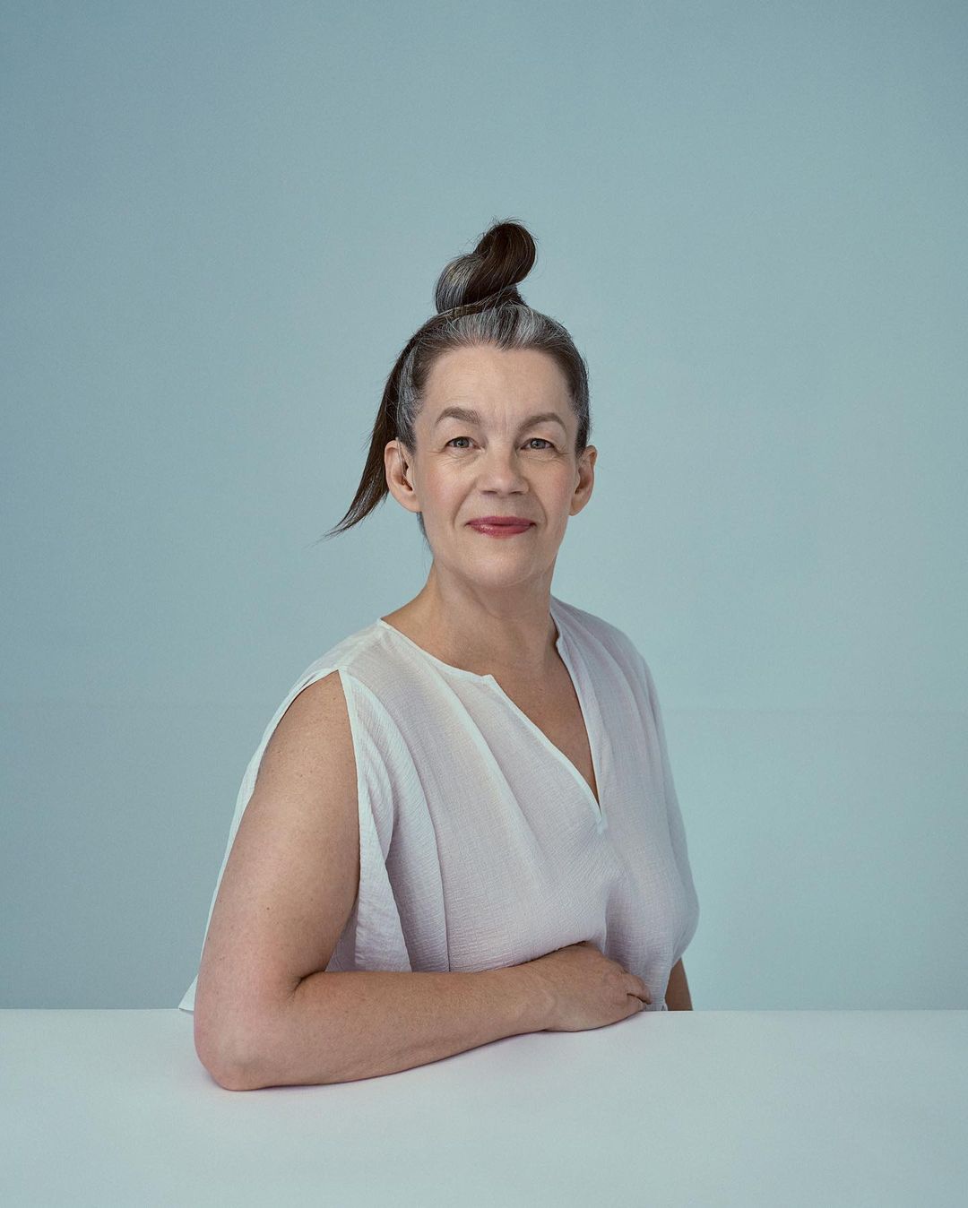 Portrait of a mature woman with gray hair styled in a topknot, wearing a white blouse, posing indoors against a light blue background.