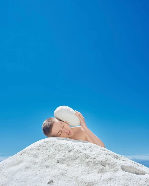 A person with short hair rests on a pile of white rocks, holding a large white rock against their head under a clear blue sky.