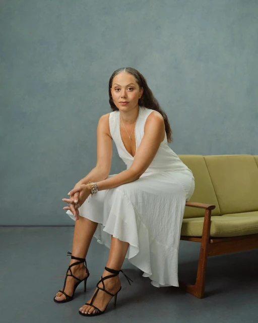 Attractive adult woman poses sitting on a sofa in a white dress and black heels in a studio with a grey backdrop.