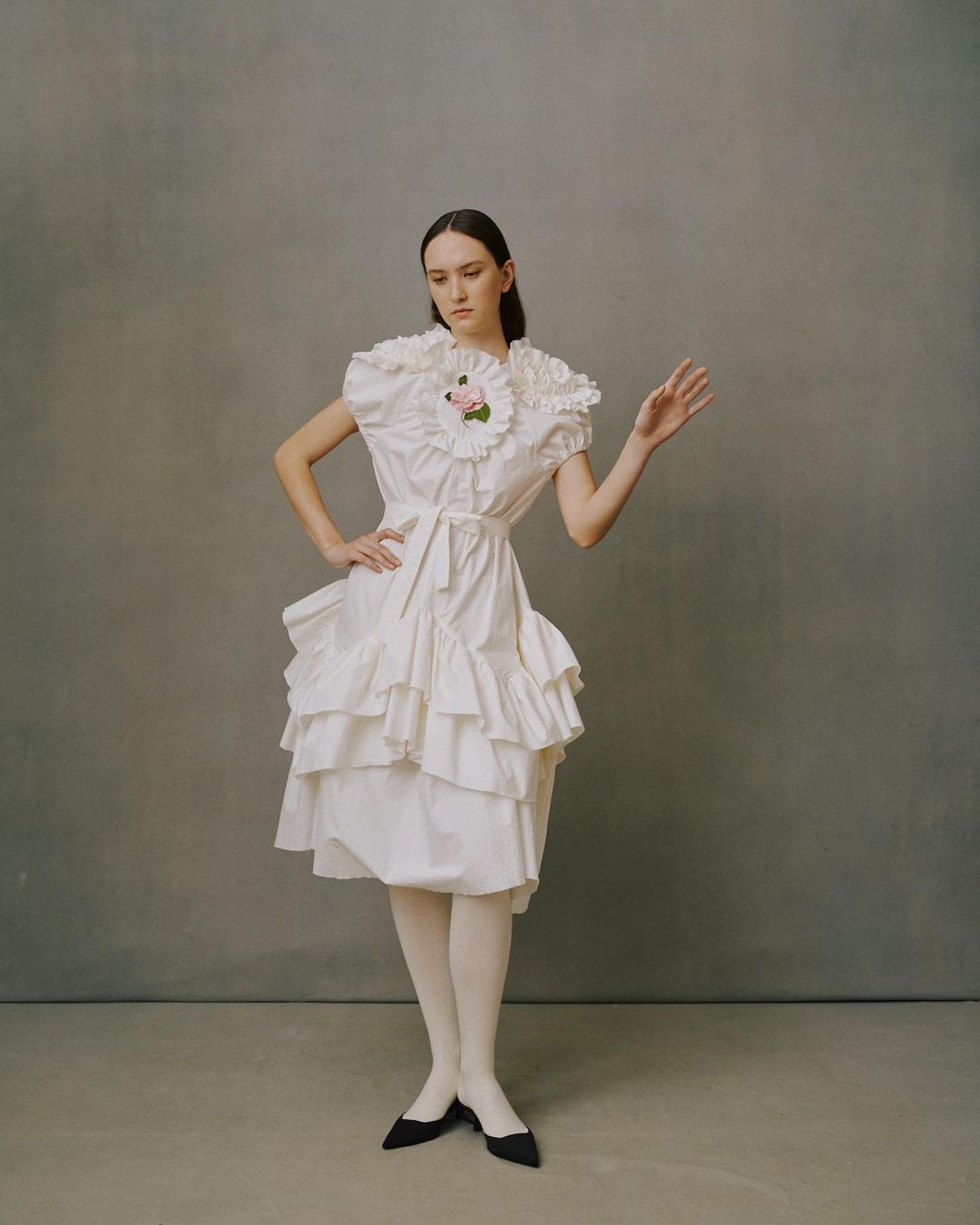 Young woman in a vintage-inspired, off-white, ruffled dress with white tights and black pointy-toe shoes stands against a grey backdrop.
