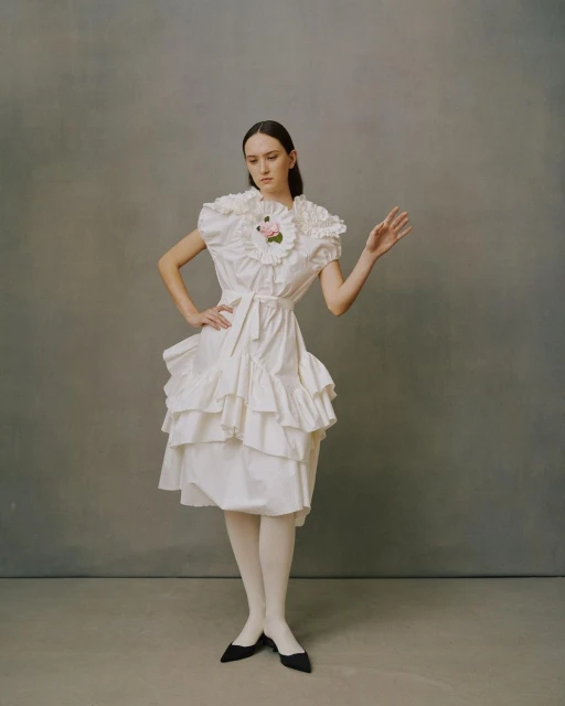 Young woman in a vintage-inspired, off-white, ruffled dress with white tights and black pointy-toe shoes stands against a grey backdrop.