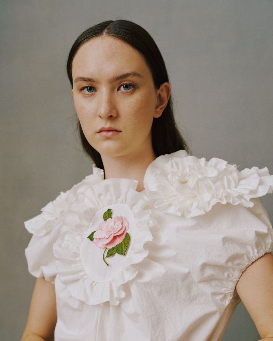 Portrait of a young woman with dark hair in a white ruffled collar top with a pink rose embroidery, looking at the camera.