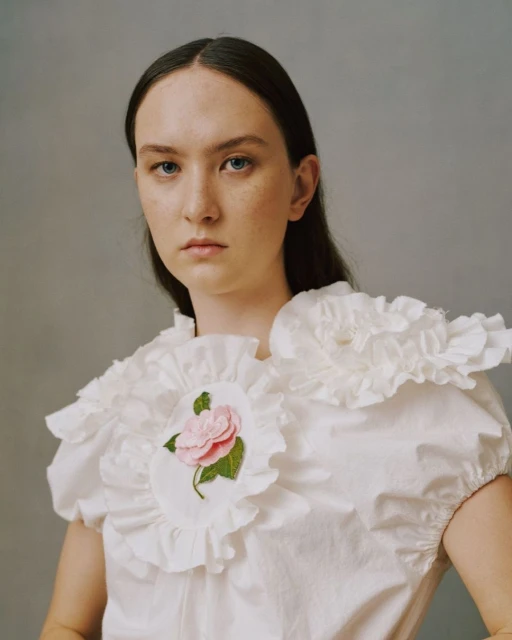 Portrait of a young woman with dark hair in a white ruffled collar top with a pink rose embroidery, looking at the camera.
