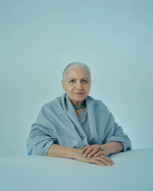 Portrait of an elderly woman with silver hair wearing a blue striped top and a green beaded necklace, posed in front of a light blue backdrop.
