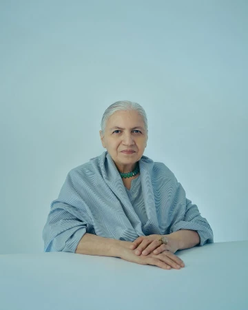 Portrait of an elderly woman with silver hair wearing a blue striped top and a green beaded necklace, posed in front of a light blue backdrop.