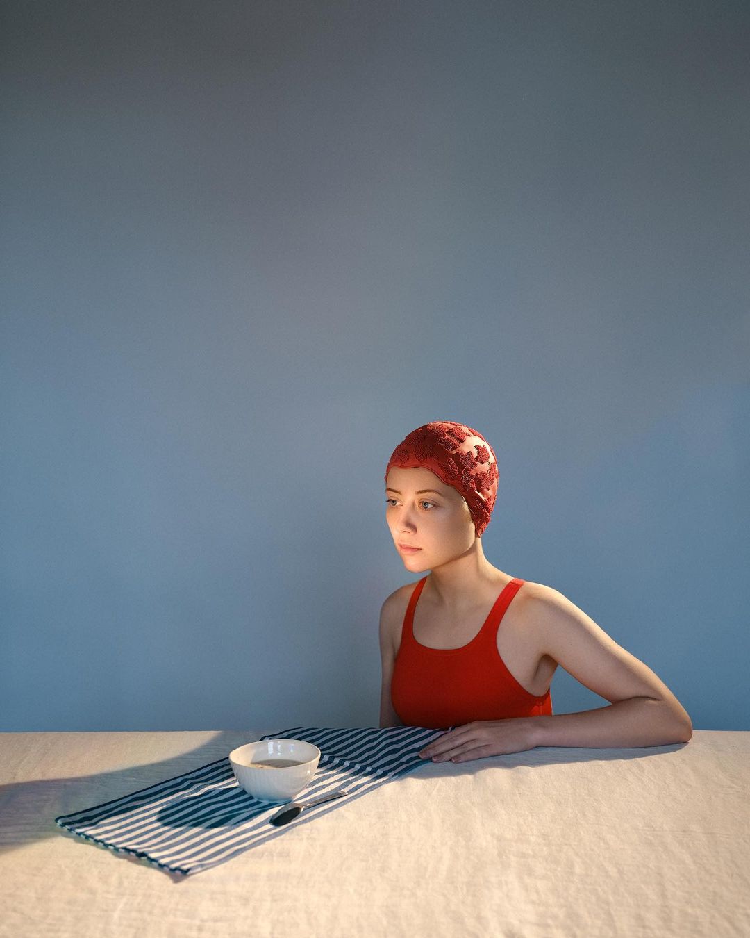 Woman in red swimsuit and swim cap seated at a table with a bowl and striped placemat against a blue background in studio lighting.